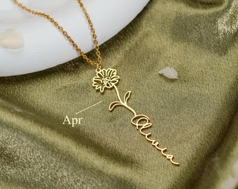 Könnte beinhalten: Eine goldfarbene Halskette mit einer zarten Kette und einem Anhänger in Blumenform, dessen Stiel in den in Schreibschrift geschriebenen Namen Olivia übergeht. Der Text Apr ist in kleinen Buchstaben neben dem Blumenstiel abgebildet.