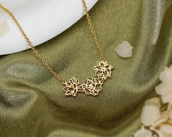 Könnte beinhalten: Eine goldfarbene Halskette mit einer feinen Kette und einem Anhänger aus drei miteinander verbundenen, durchbrochenen Blumenmotiven. Jedes Blumendesign ist filigran gearbeitet und mit einem kleinen farbigen Stein in der Mitte versehen.