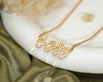 Könnte beinhalten: Eine goldfarbene Halskette mit einem Anhänger, der das Wort LOVE darstellt, buchstabiert durch vier verschiedene Handzeichen der amerikanischen Gebärdensprache. Die feine Kette ist an beiden Enden des metallenen Handzeichen-Motivs befestigt.