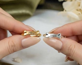 Könnte beinhalten: Zwei Ringe mit einem hundeknochenförmigen Mittelstück. Ein Ring ist goldfarben mit dem eingravierten Namen Olivia auf dem Knochen, der andere ist silberfarben mit dem eingravierten Namen Victoria. Beide Ringe haben eine dünne, polierte Ringschiene.