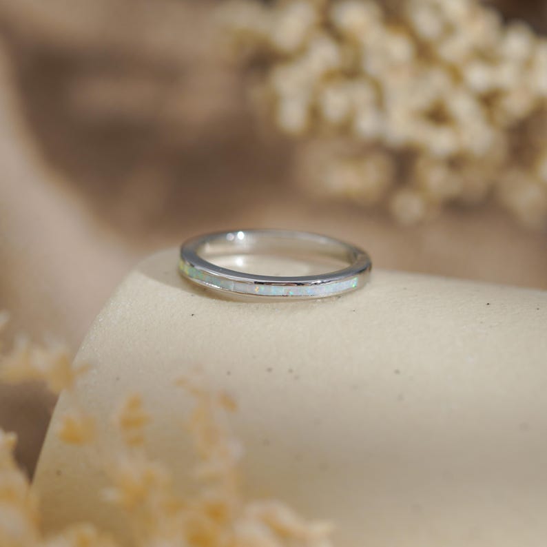 May include: A silver ring with a band of white opal stones. The ring is set on a neutral-toned surface, with soft, blurred details in the background. The ring is a delicate piece of jewelry.