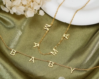 Könnte beinhalten: Zwei goldfarbene Halsketten mit dünnen Metallketten. Eine Halskette zeigt die Buchstaben M-A-M-A, die entlang der Kette verteilt sind, während die zweite Halskette die Buchstaben B-A-B-Y in das Kettendesign integriert.