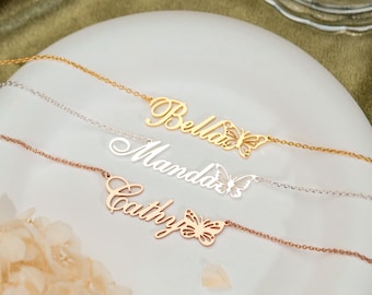 Könnte beinhalten: Drei zarte Halsketten mit kursiven Namensschildern und Schmetterlingsanhängern. Die oberste Kette ist goldfarben mit dem Namen Bella, die mittlere ist silberfarben mit Manda und die untere ist roségoldfarben mit dem Namen Cathy.
