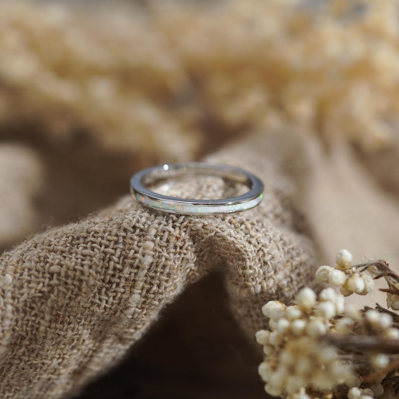 May include: A silver ring with a white opal band. The ring is set against a neutral-toned, textured background, with soft, blurred elements in the background. The ring is a delicate piece of jewelry.