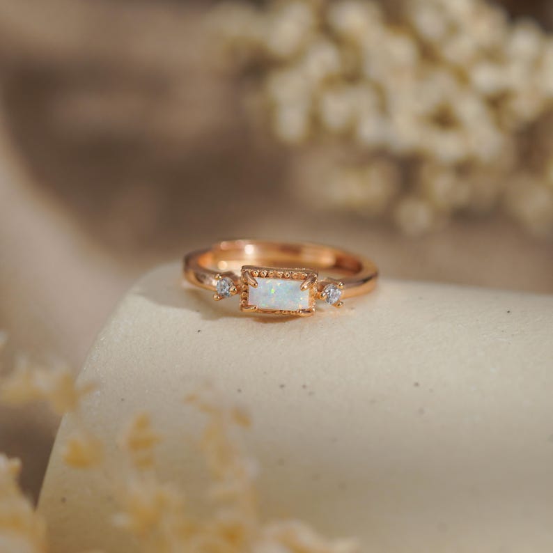 May include: Rose gold ring featuring a rectangular opal center stone and two smaller accent stones. The ring has a delicate, detailed setting and a polished finish. The opal has a milky white appearance, and the ring is set on a neutral surface.