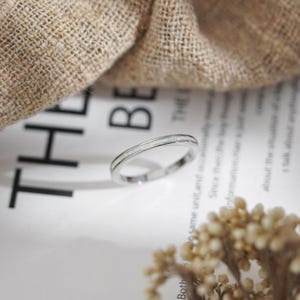 May include: A silver ring with a thin band and a white opal inlay. The ring is set against a backdrop of a white paper with text and a beige fabric.
