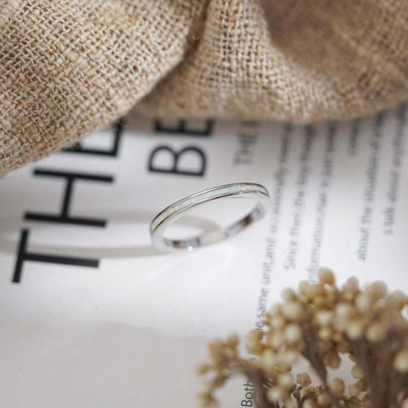 May include: A silver ring with a thin band and a white opal inlay. The ring is set against a backdrop of a white paper with text and a beige fabric.