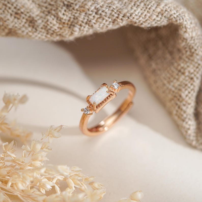 May include: Rose gold adjustable ring featuring a rectangular opal center stone and two smaller accent stones. The ring has a delicate, minimalist design, perfect for everyday wear. The opal has a milky white appearance.