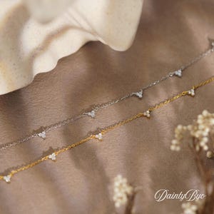 Puede incluir: Dos delicados collares, uno plateado y otro dorado, cada uno adornado con pequeños y brillantes grupos triangulares de gemas. Los collares se exhiben sobre una superficie neutra y texturizada, con la marca "DaintyByo".