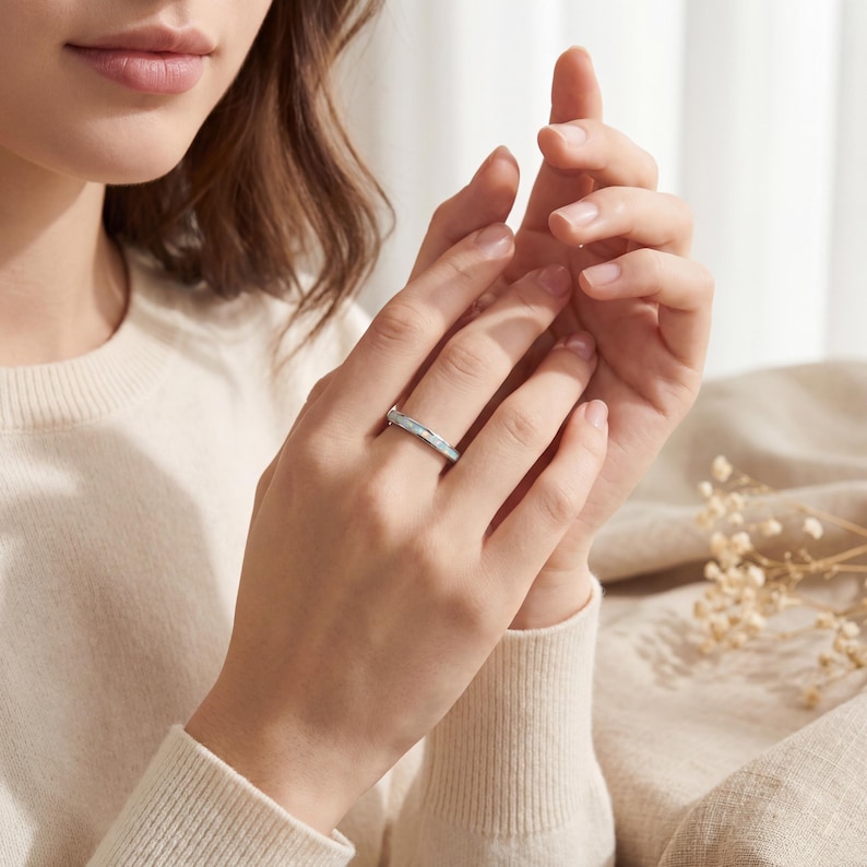 May include: A silver ring with a textured band and a light-colored inlay. The ring is worn on a finger, showcasing its design and details. The jewelry piece is a simple yet elegant accessory.