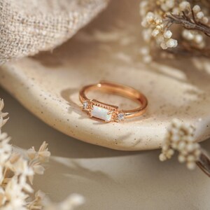 Puede incluir: Anillo ajustable de oro rosa con una piedra central de &oacute;palo blanco rectangular y dos peque&ntilde;as piedras de acento transparentes. El anillo est&aacute; sobre una superficie de tonos neutros con flores secas en el fondo. Joyer&iacute;a.
