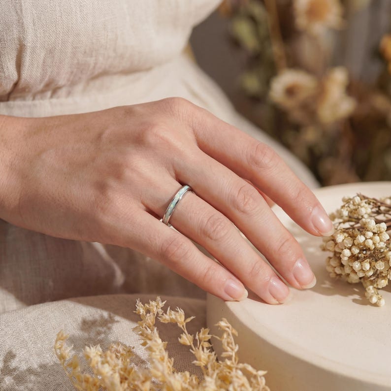 May include: A silver ring with a white inlay is displayed on a person's finger. The ring has a thin band and a textured surface. The background includes a beige fabric and dried flowers, creating a soft, natural aesthetic.