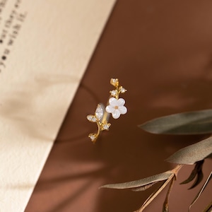 Puede incluir: Un anillo floral dorado con una flor blanca y cristales transparentes. El anillo tiene un diseño delicado inspirado en la naturaleza con una estructura similar a una rama. El anillo está sobre un fondo marrón y beige.