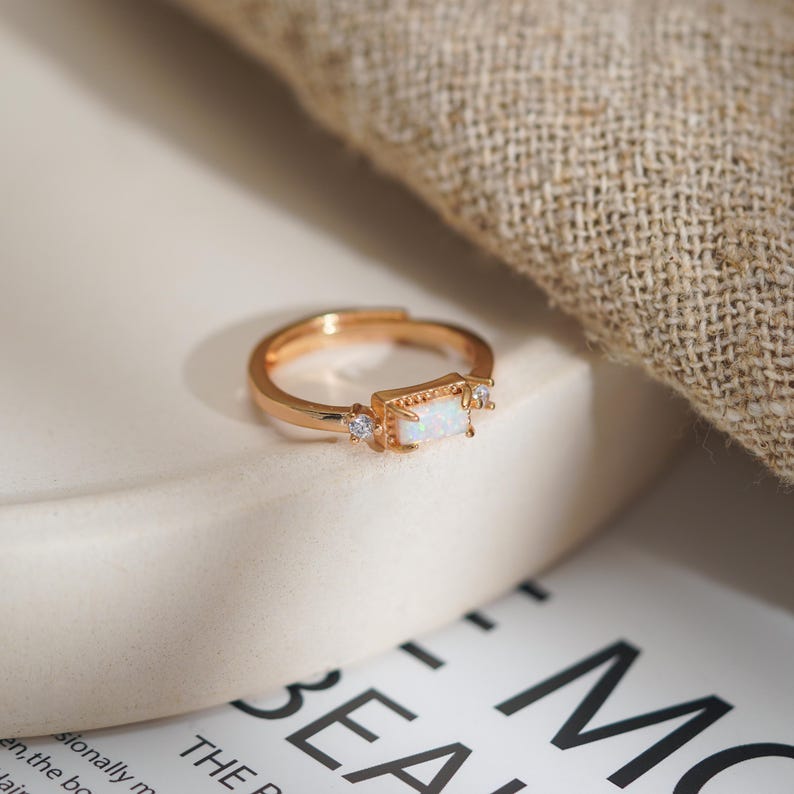 Puede incluir: Un anillo ajustable dorado con una piedra central de &oacute;palo rectangular y peque&ntilde;as piedras de acento transparentes. El anillo est&aacute; sobre una superficie blanca con un fondo borroso.