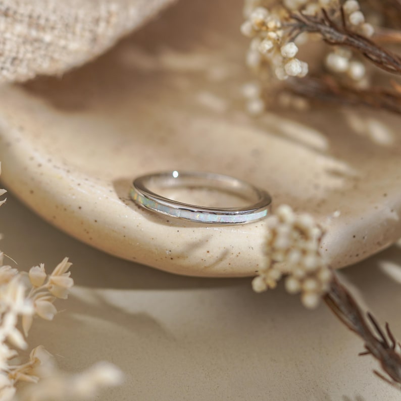 May include: A silver ring with a band of white opal. The ring is set on a beige, textured surface, with dried flowers in the background. The ring is a delicate piece of jewelry.