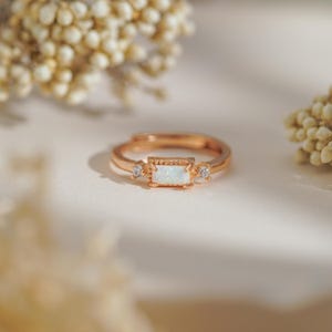 Puede incluir: Anillo ajustable de oro rosa con una piedra central de &oacute;palo rectangular y dos piedras redondas m&aacute;s peque&ntilde;as. El anillo tiene un dise&ntilde;o delicado con una banda texturizada. El anillo est&aacute; sobre un fondo suave y neutro.