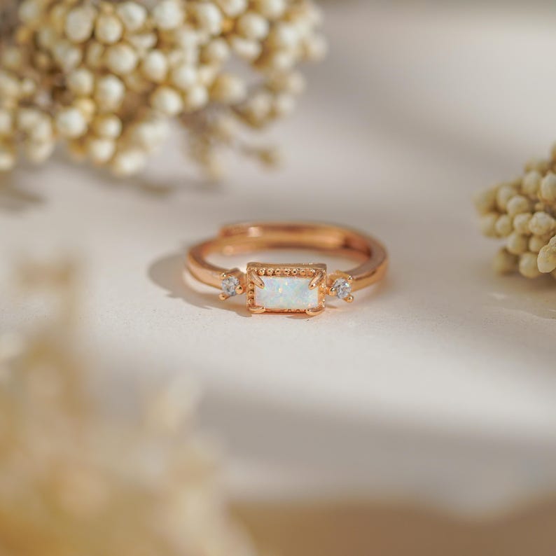 Puede incluir: Anillo ajustable de oro rosa con una piedra central de &oacute;palo rectangular y dos piedras redondas m&aacute;s peque&ntilde;as. El anillo tiene un dise&ntilde;o delicado con una banda texturizada. El anillo est&aacute; sobre un fondo suave y neutro.