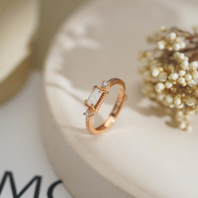 Puede incluir: Un anillo en tono oro rosa con una piedra rectangular blanca de aspecto opalino engastada horizontalmente en el centro, flanqueada por dos peque&ntilde;as piedras redondas transparentes a cada lado, con una textura met&aacute;lica perlada.