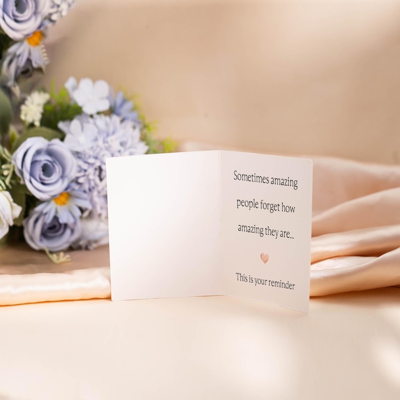May include: A white greeting card with the text "Sometimes amazing people forget how amazing they are... This is your reminder" and a small pink heart. A bouquet of blue and white flowers is in the background.