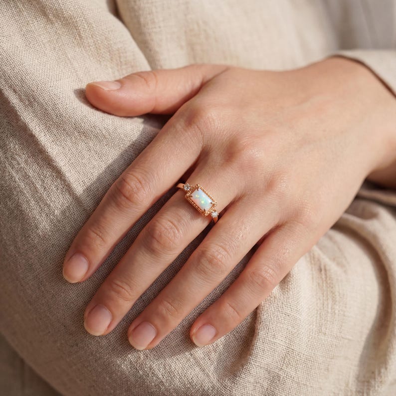 Puede incluir: Anillo de oro rosa con una piedra preciosa de &oacute;palo rectangular. El anillo tiene un dise&ntilde;o ornamentado con peque&ntilde;as piedras de acento. El anillo se lleva en una mano, destacando sus elegantes detalles y artesan&iacute;a. El &oacute;palo es el punto focal.