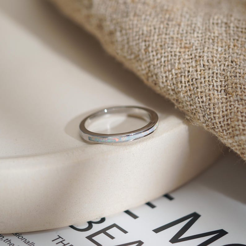 May include: A silver ring with a thin band and a white opal inlay. The opal has subtle pink and orange hues. The ring is set on a white surface with a neutral-colored fabric in the background. The ring is a piece of jewelry.