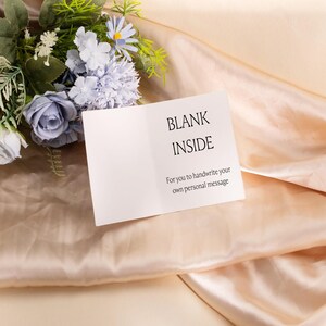 May include: A blank greeting card with the words "BLANK INSIDE" printed on the inside, accompanied by a floral arrangement of blue and white flowers. The card is on a peach-colored satin fabric.