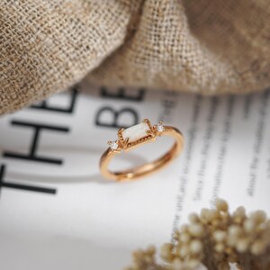 Puede incluir: Un anillo dorado con una piedra central de &oacute;palo blanco rectangular y dos peque&ntilde;as piedras claras de acento. El anillo est&aacute; sobre una superficie blanca con texto borroso y una tela beige en el fondo.