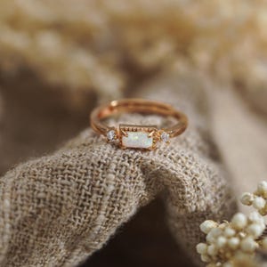 Puede incluir: Un anillo en tono oro rosa con una piedra de &oacute;palo rectangular central engastada en un marco de cuentas, flanqueada por dos peque&ntilde;as piedras redondas transparentes a cada lado. La banda es fina y met&aacute;lica con acabado pulido.