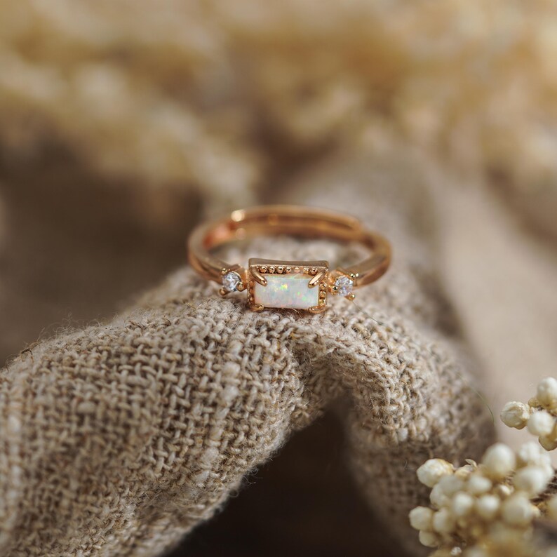 Puede incluir: Un anillo en tono oro rosa con una piedra de &oacute;palo rectangular central engastada en un marco de cuentas, flanqueada por dos peque&ntilde;as piedras redondas transparentes a cada lado. La banda es fina y met&aacute;lica con acabado pulido.