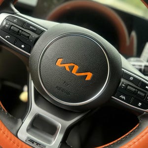 May include: A car steering wheel with a black centre and orange trim. The centre displays an orange Kia logo and the word "AIRBAG". The steering wheel has buttons for mode and other functions.