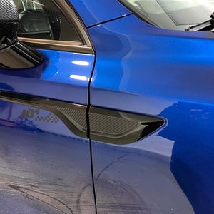 Pode incluir: Vista de perto da carroçaria azul vibrante de um carro, com um detalhe preto com textura de fibra de carbono. O espelho retrovisor é visível. O texto "JB4" e um desenho de bandeira quadriculada também estão presentes.