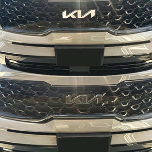 Puede incluir: Primer plano de la parrilla delantera de un coche, con el logotipo de Kia en plata. La parrilla tiene un patrón de panal de abeja negro con un borde plateado. Una placa rectangular negra está montada en la parrilla.