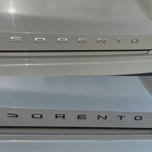 Puede incluir: Primer plano de la parte trasera de un coche plateado con las letras cromadas "SORENTO". Las letras están montadas individualmente sobre un panel plateado, reflejando la luz. La carrocería del coche es de color plateado claro.