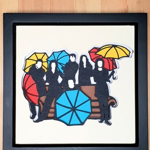 May include: A framed papercraft artwork featuring six silhouettes of people sitting on a brown couch with colorful umbrellas above them. The artwork is on a white background.