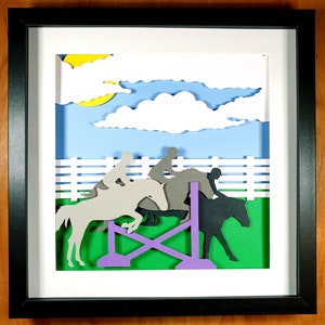 May include: A framed paper art piece depicting a show jumping scene. Silhouettes of horses and riders are shown jumping over a purple and green obstacle. The background features a blue sky with white clouds and a yellow sun.