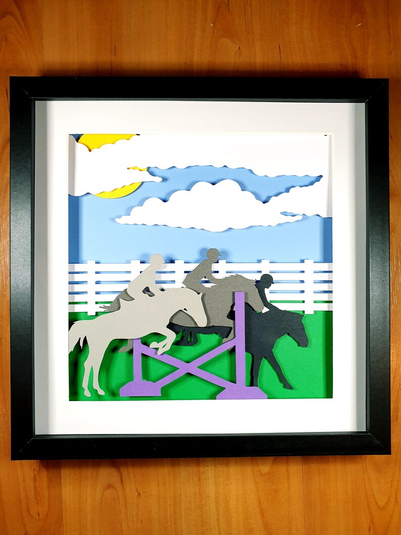 May include: A framed paper art piece depicting a show jumping scene. Silhouettes of horses and riders are shown jumping over a purple and green obstacle. The background features a blue sky with white clouds and a yellow sun.