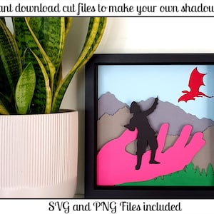 May include: A shadow box with a black frame and a pink hand holding a silhouette of a person. The person is pointing at a red dragon flying in a blue sky. The background includes mountains and green trees. The text "SVG and PNG Files included" is at the bottom of the image.