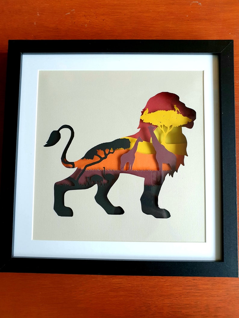 May include: A paper cut-out of a lion with a sunset and African savanna scene inside the silhouette. The lion is black, the sunset is orange, yellow, and red, and the savanna is green and brown.