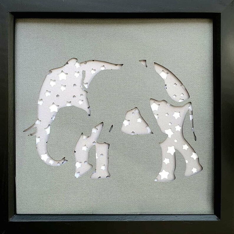 May include: A grey framed picture with a white silhouette of two elephants cut out of the background. The elephants are covered in white stars.