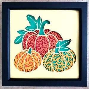 Pu&ograve; includere: Un'opera d'arte in carta incorniciata con tre zucche con intricati disegni ritagliati. Le zucche sono sovrapposte, con la zucca pi&ugrave; grande sul retro e la zucca pi&ugrave; piccola sul davanti. Le zucche sono in tonalit&agrave; di arancione, rosso e verde.