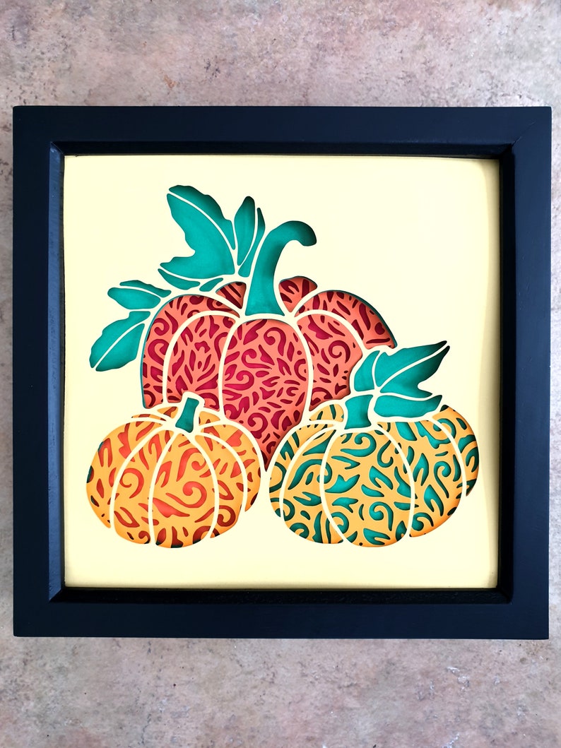 Pu&ograve; includere: Un'opera d'arte in carta incorniciata con tre zucche con intricati disegni ritagliati. Le zucche sono sovrapposte, con la zucca pi&ugrave; grande sul retro e la zucca pi&ugrave; piccola sul davanti. Le zucche sono in tonalit&agrave; di arancione, rosso e verde.