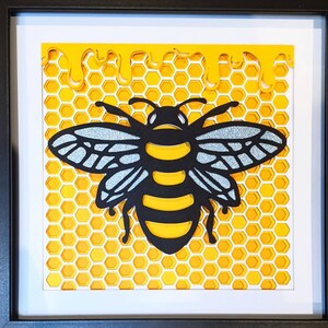 Puede incluir: Una abeja recortada en papel negro y amarillo con alas de purpurina plateada se coloca sobre un fondo de panal amarillo. El panal gotea pintura amarilla.