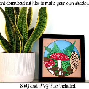 May include: A shadow box craft project featuring a red and white mushroom, brown mushrooms, and green ferns. The shadow box is framed in black and has a light blue background. The text "SVG and PNG Files included" is at the bottom of the image.
