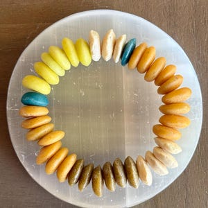 Op de afbeelding: Een kralenarmband met een kleurengradatie van geel naar oranje, met enkele turquoise en gebroken witte kralen. De armband wordt gepresenteerd op een rond, doorschijnend, wit oppervlak.