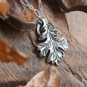 May include: A silver necklace with a detailed oak leaf pendant. The necklace is displayed on a wooden surface with dried leaves.