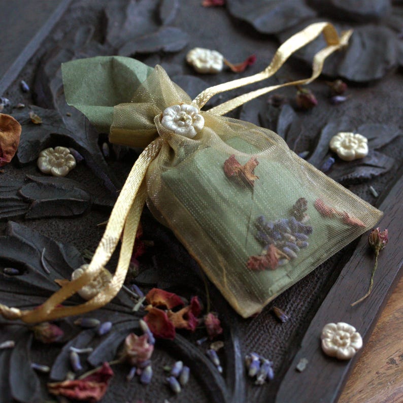 May include: A small, green and gold sachet filled with dried flowers and herbs. The sachet is tied with a gold ribbon and is sitting on a dark wooden surface.