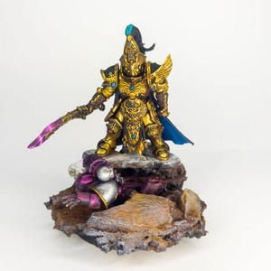 Peut inclure: Une figurine miniature peinte en or d'un guerrier debout sur une base rocheuse. Le guerrier tient une épée et porte une cape bleue. La figurine est peinte avec des détails complexes et est un excellent exemple de peinture miniature.