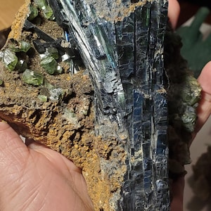 May include: A hand holds a mineral specimen featuring a large, dark, metallic-looking crystal formation. The crystal has a layered, columnar structure with a deep blue-green iridescence. Smaller, green crystal clusters are also visible.
