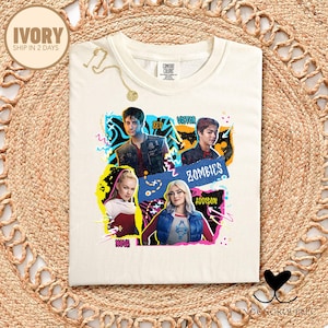 May include: Ivory-colored t-shirt featuring a colorful graphic of characters from the movie "Zombies". The design includes the names Zed, Victor, Addison, and Nova. The shirt has a round neckline and short sleeves.