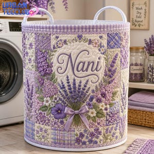 May include: A cylindrical quilted laundry basket featuring a lavender and floral patchwork pattern. The front displays the name Nani in cursive script above a bouquet of lavender tied with a purple ribbon. It has two white top handles.